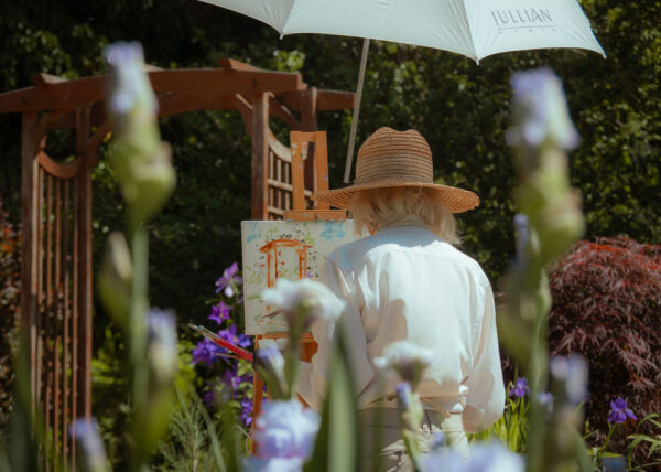 photo of a woman with white hair, wearing a straw hat, painting on an easel in a garden