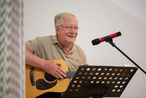 Image of an older gentleman playing an acoustic guitar while smiling on open mic night